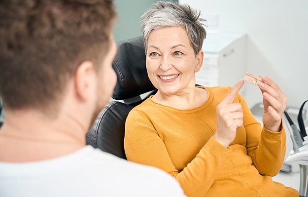 Woman in yellow shirt in dental chair holding and pointing to sample dentures