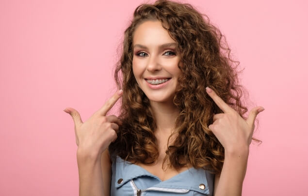 Woman with curly hair and braces pointing to her teeth