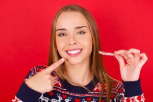 Woman in red and blue holiday sweater pointing to her teeth and smiling holding up her Invisalign